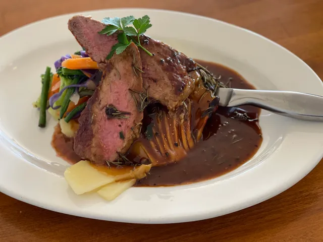 Sliced roast lamb with dark jus, potato gratin and colourful vegetables on white oval plate