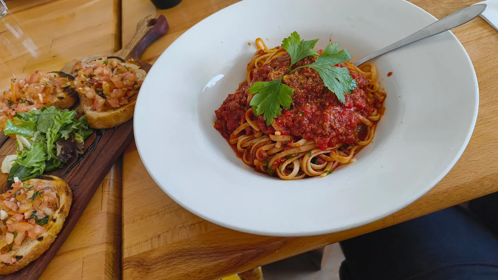 Spaghetti bolognese with parsley in white bowl alongside bruschetta board with tomato salsa