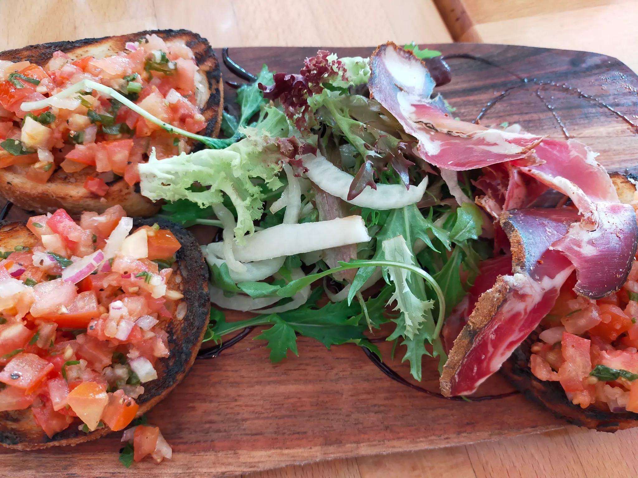 Wooden board with bruschetta topped with fresh tomato salsa, mixed greens and sliced prosciutto